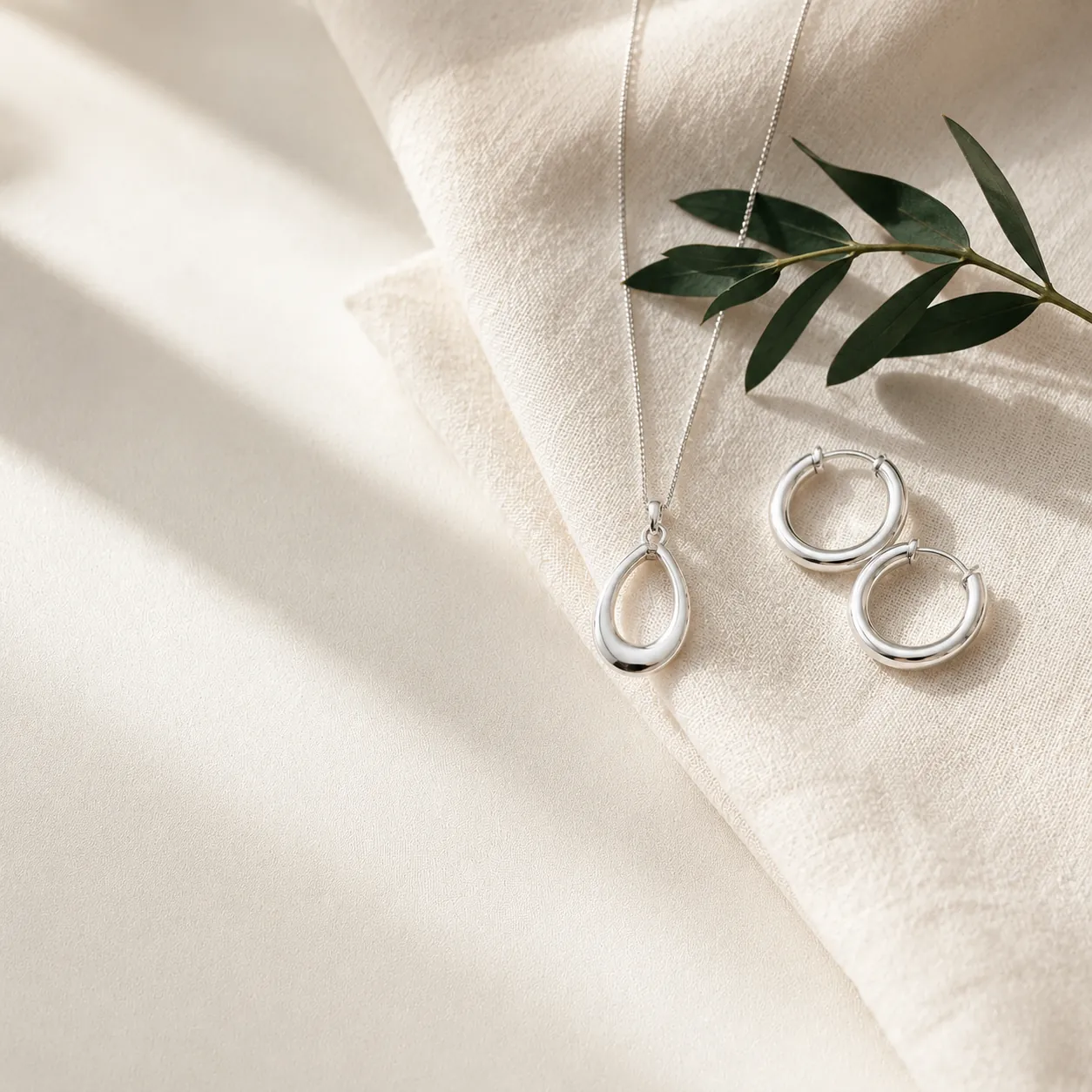 A silver pendant and hoops on folded ivory linen with an emerald leaf sprig